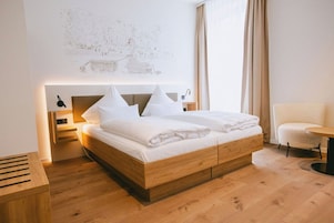 A modern hotel room with a wooden bed, a small bedside table, a chair, and a wall-mounted artwork.
