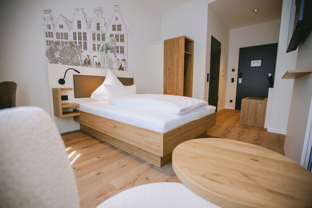 A modern hotel room with a wooden bed, a bedside table, a lamp, and a wall-mounted artwork.