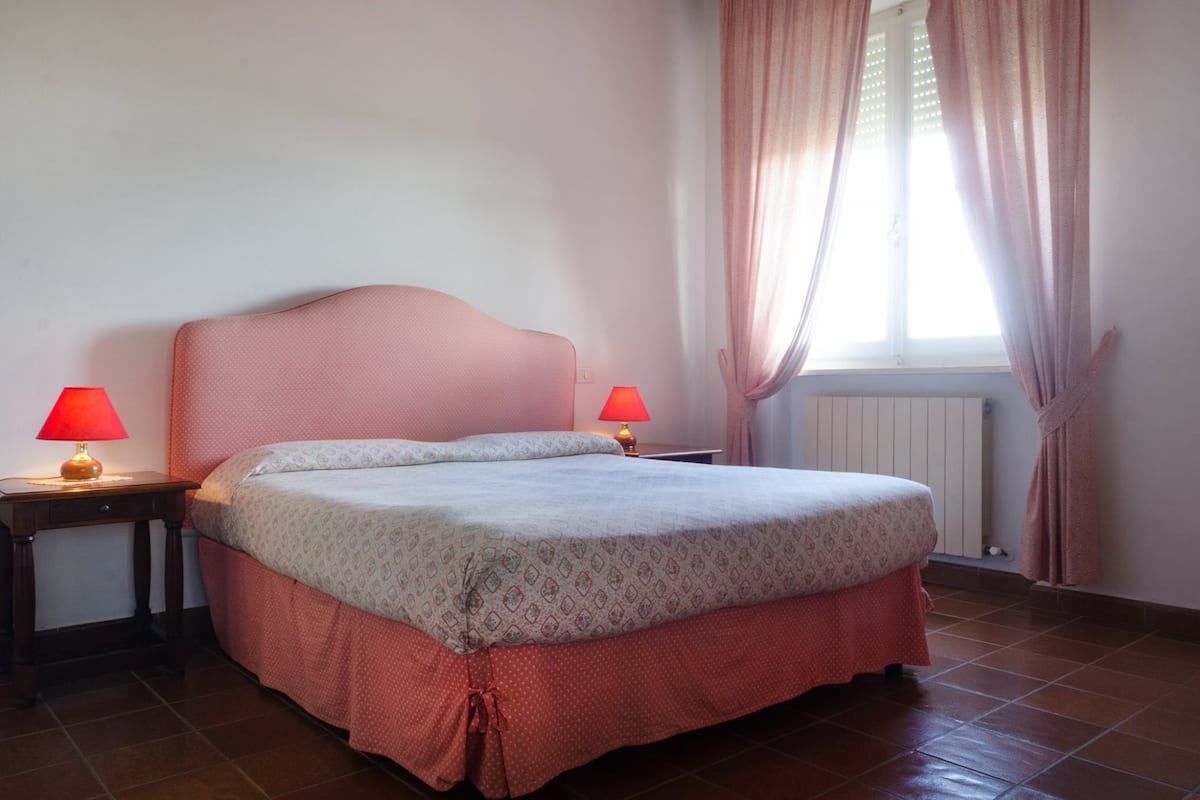 Une chambre à coucher avec un lit, deux tables de chevet avec des lampes, une fenêtre avec des rideaux et un radiateur.