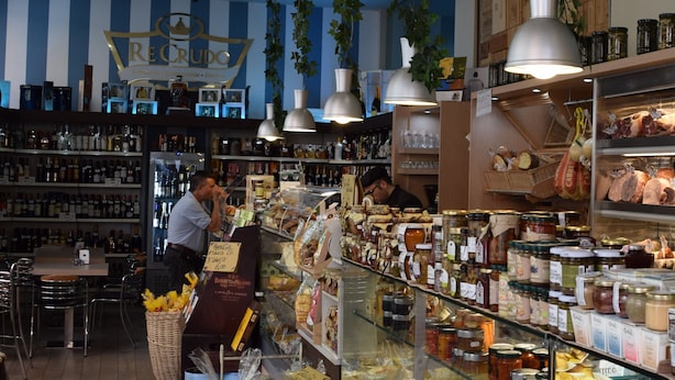 Een winkelinterieur met schappen vol diverse etenswaren en een man die bij de toonbank staat.