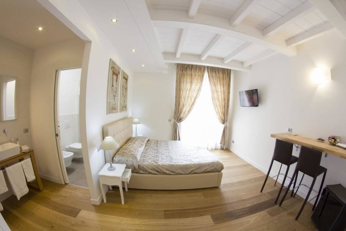 A bedroom with a bed, bedside table, and a desk with two chairs. There is a bathroom visible through an open door.