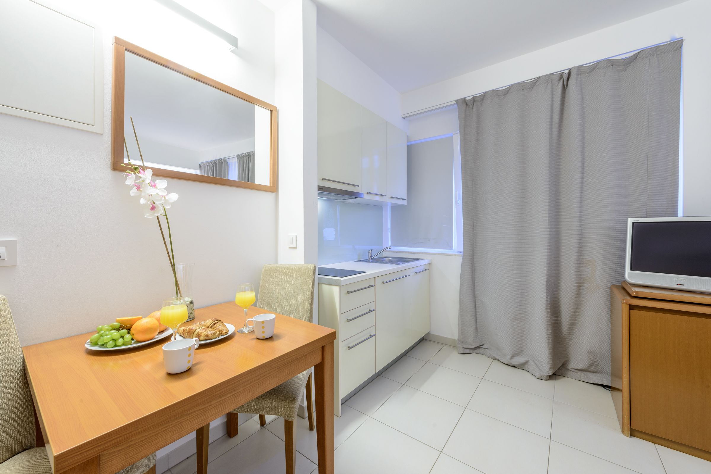 A compact kitchen with a dining area, a TV on a stand, and a window with curtains.