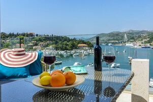 A table with a plate of oranges, a wine bottle, and a wine glass, overlooking a marina with boats and a bridge.