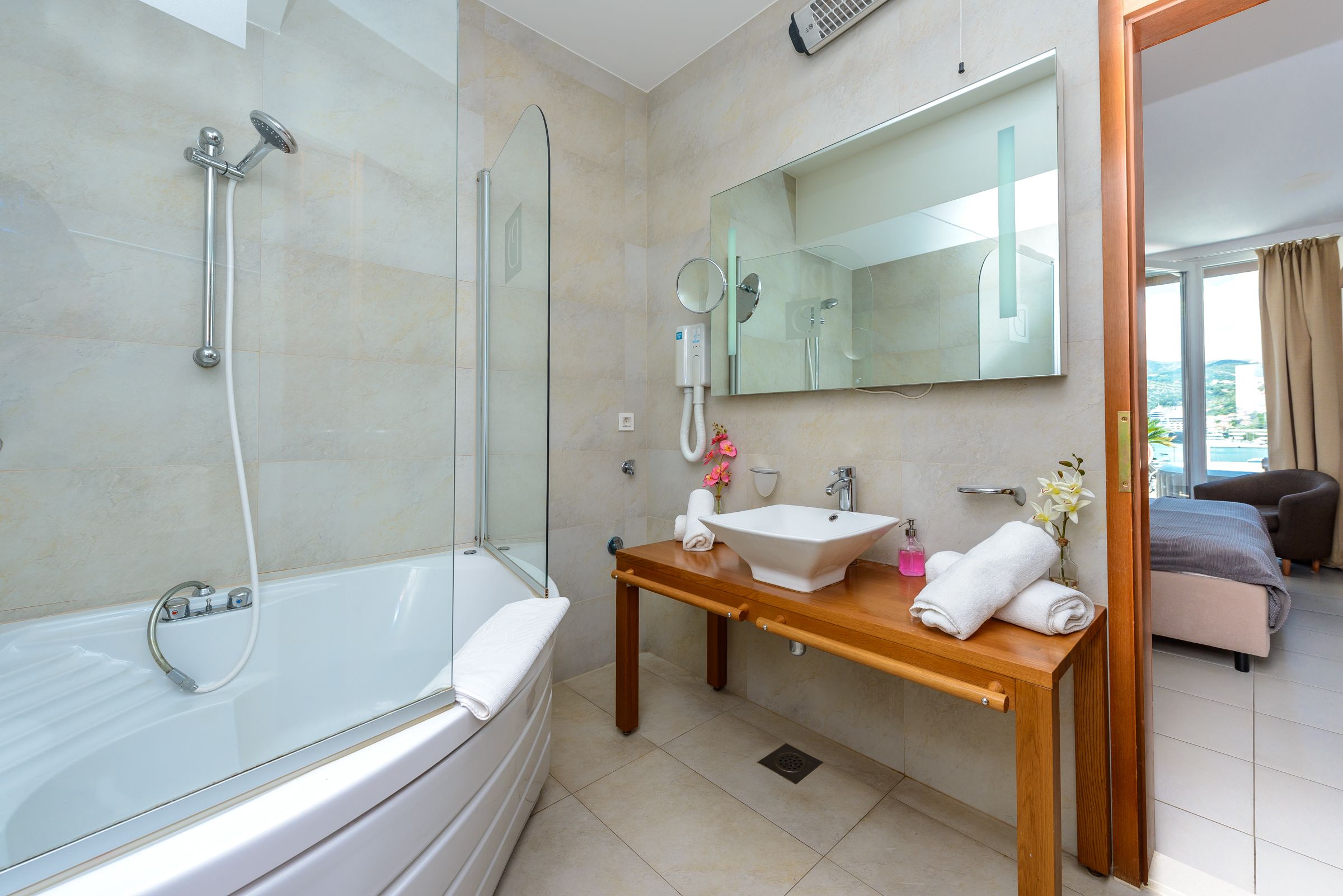 A modern bathroom with a glass-enclosed shower, a standalone bathtub, a wooden vanity with a sink and mirror, and a view of the outside through an open door.