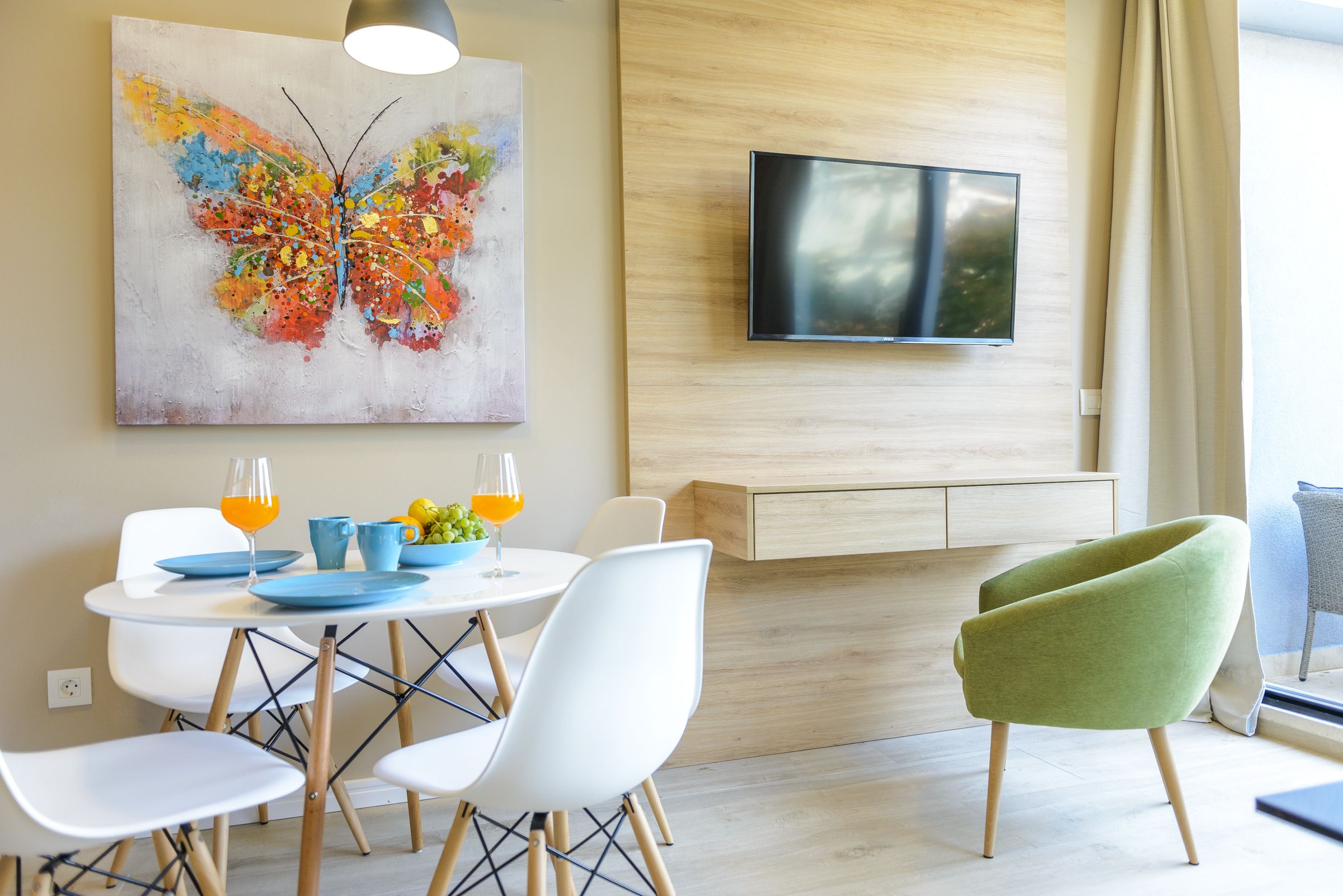 A modern dining area with a table set for two, a colorful butterfly painting, and a flat-screen TV mounted on the wall.
