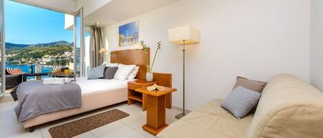 Una habitación de hotel moderna con cama, sofá, escritorio y un amplio ventanal que ofrece una vista panorámica de un pueblo costero.