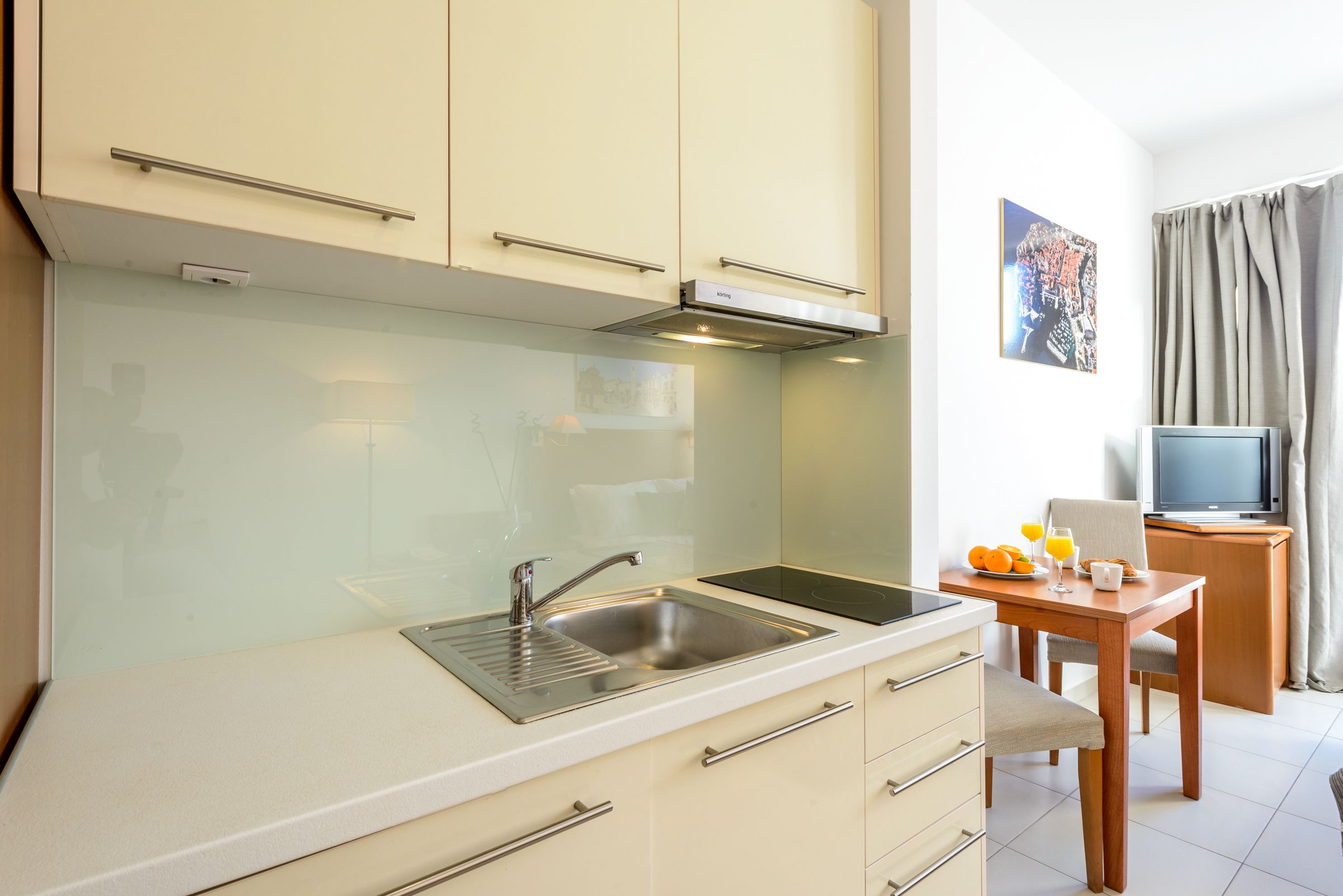 A modern kitchen with a stainless steel sink, a built-in oven, and a microwave. There is a dining table with a chair and a small television on a stand.
