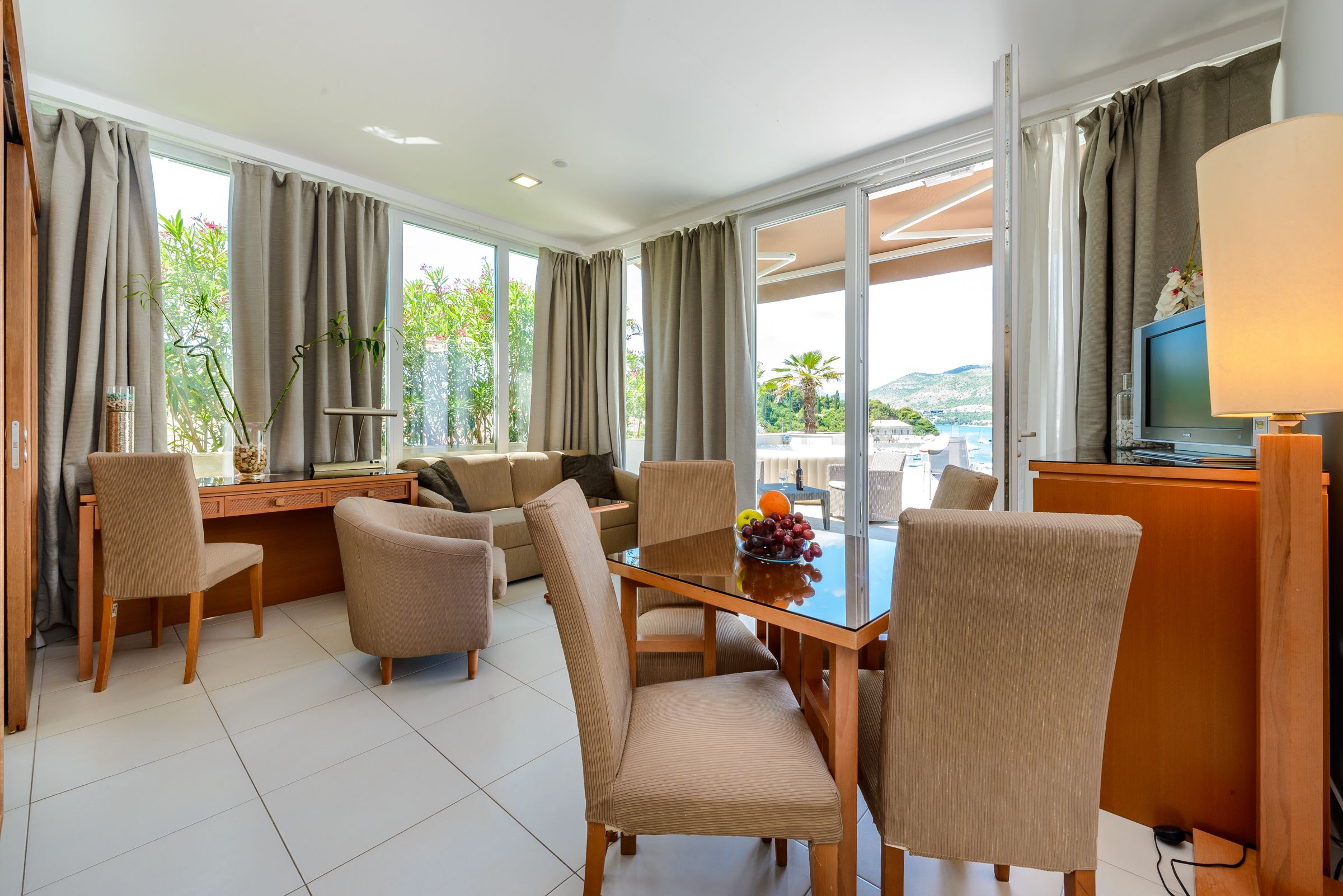 A modern hotel room with a dining area, a sofa set, a glass table with chairs, and a TV on a wooden stand.