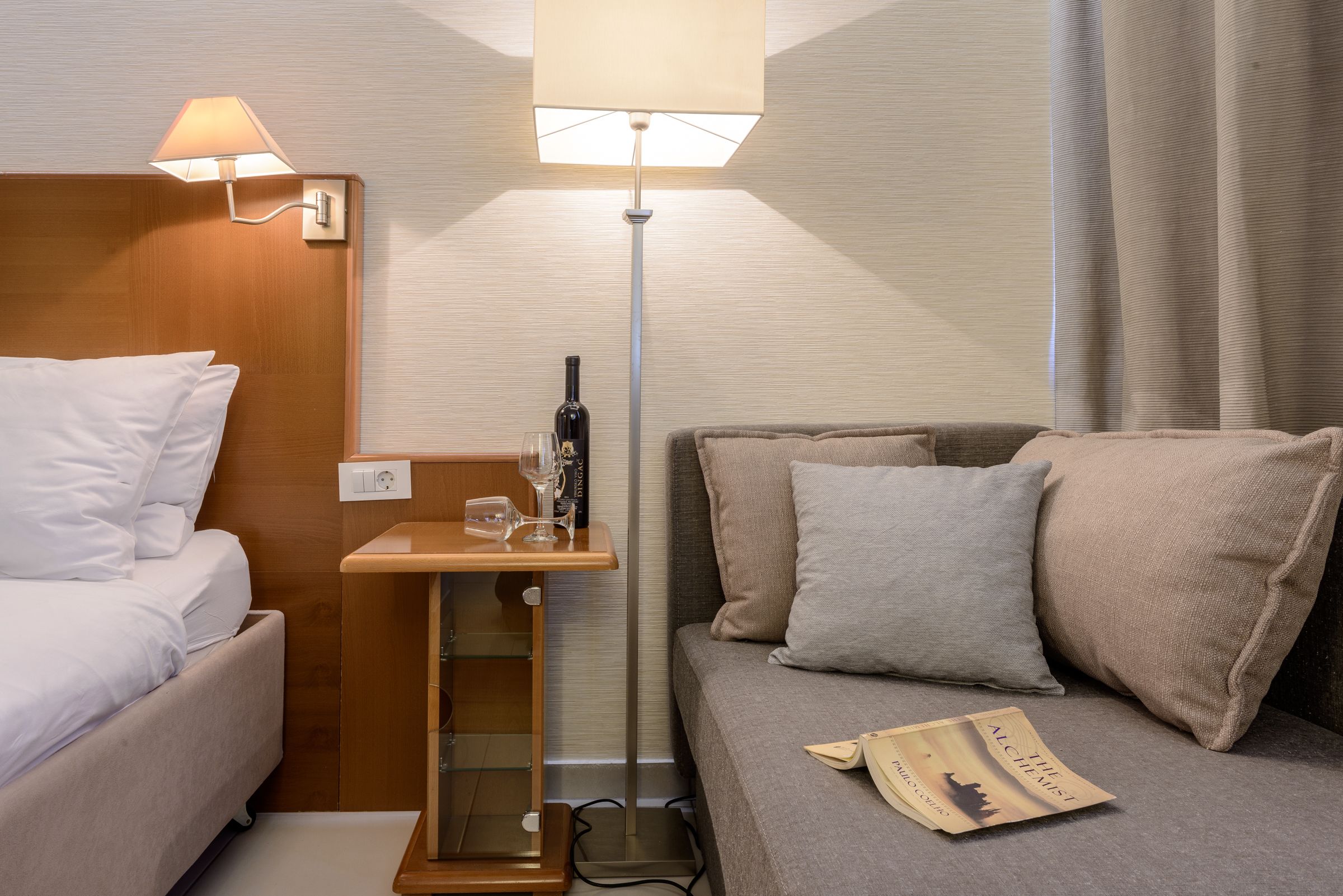 Quarto de hotel com cama, sofá, mesa de cabeceira com abajur, garrafa de vinho e um livro.
