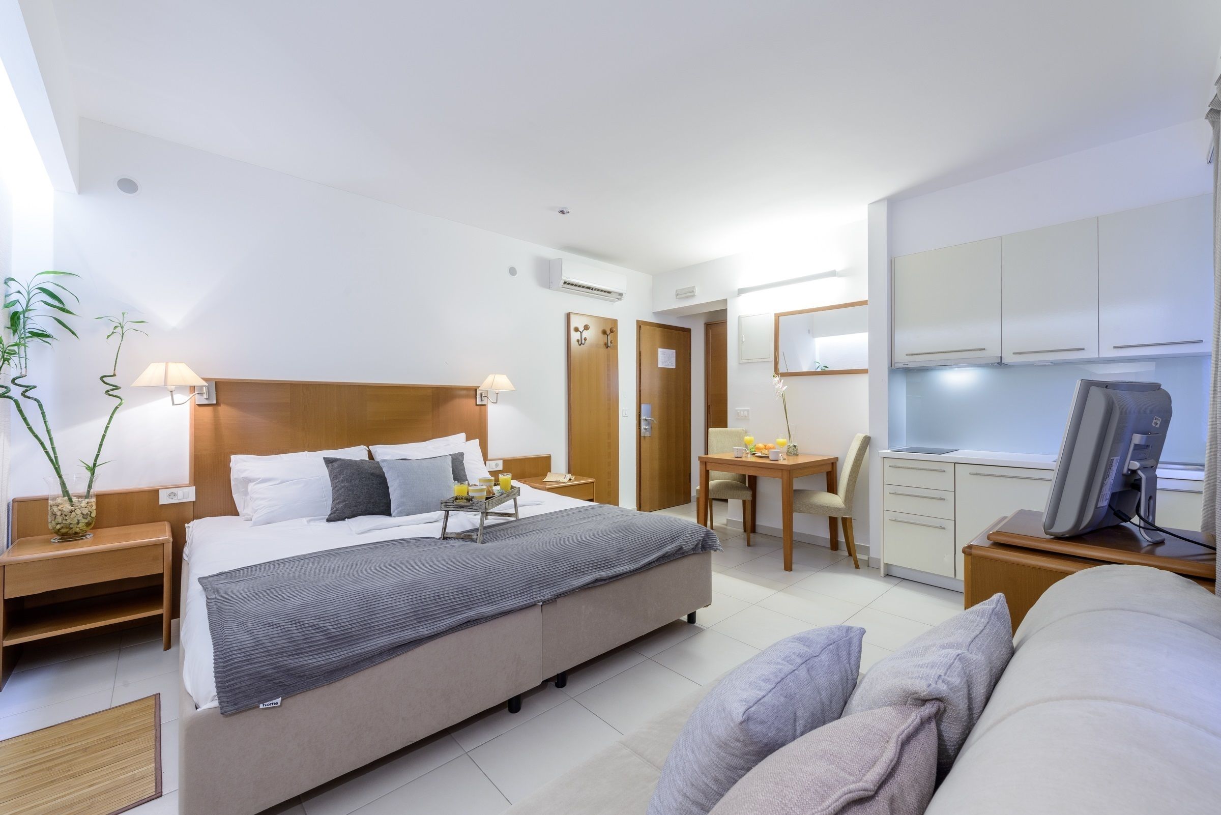 A modern hotel room with a large bed, bedside tables, a desk, a chair, a TV, and a small kitchenette.