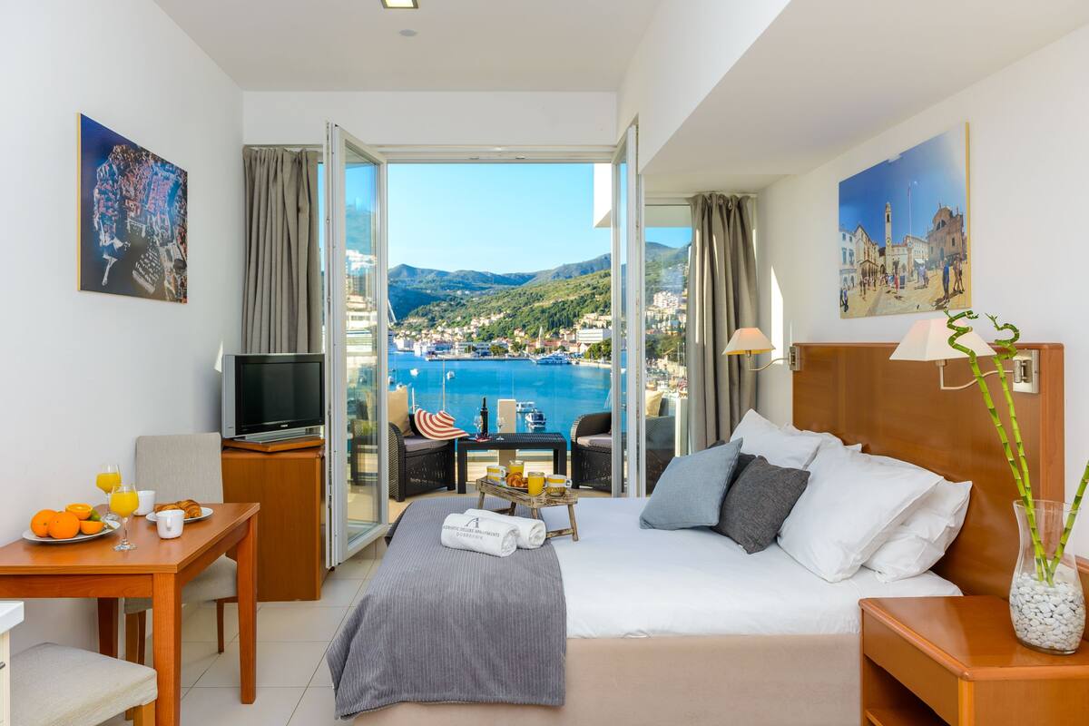 Une chambre d’hôtel avec un lit, un bureau avec un petit-déjeuner, une télévision et un balcon offrant une vue sur la mer et les montagnes.