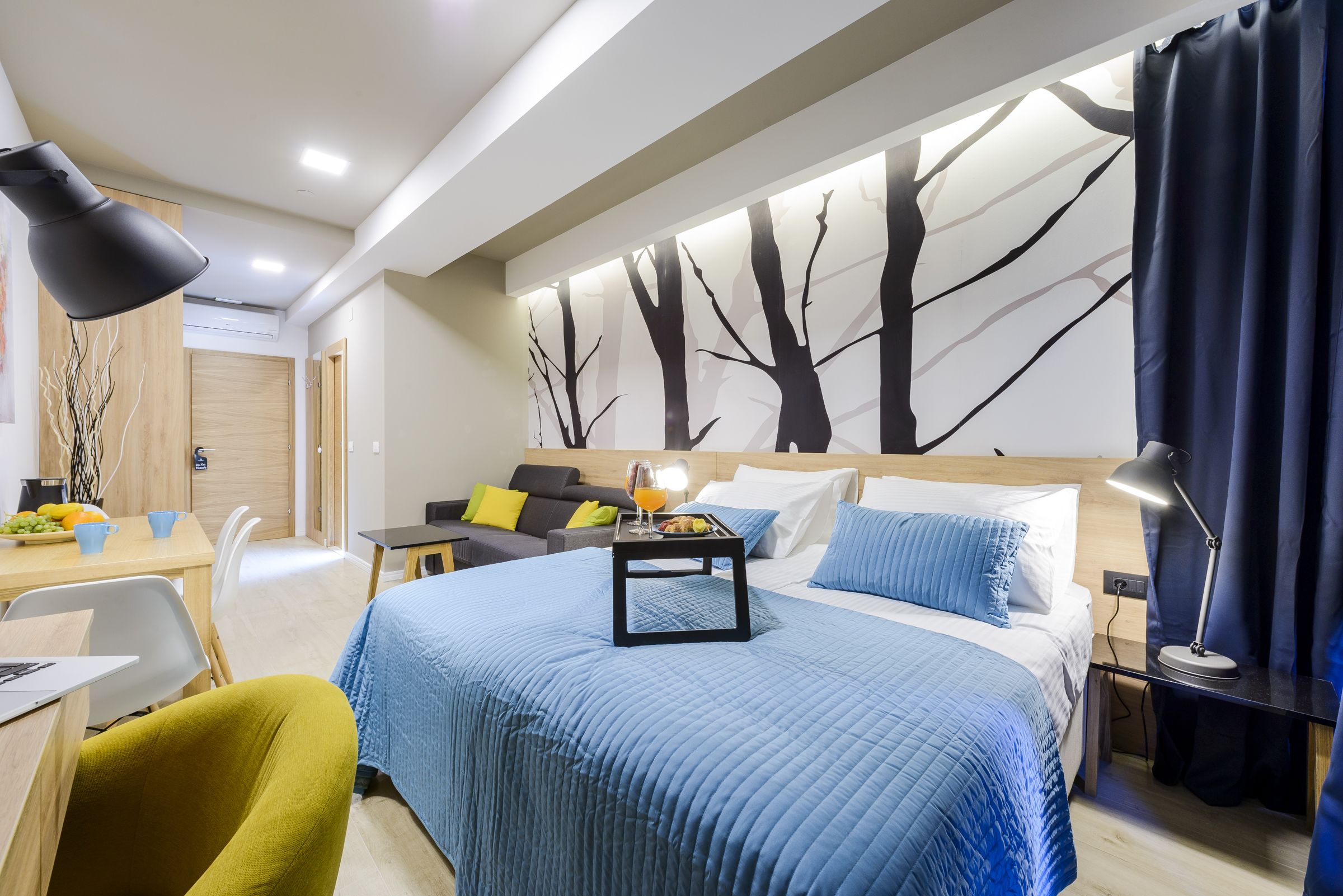 Une chambre d’hôtel moderne avec un grand lit, un bureau avec un ordinateur portable, une table à manger avec des fruits et un mur orné d’une fresque représentant un arbre.