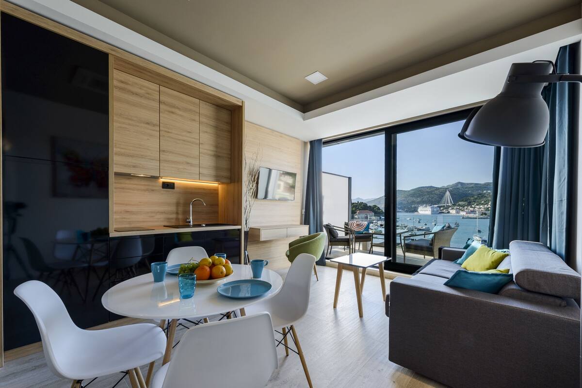 Un salon moderne avec un canapé, une table à manger et des chaises. De grandes fenêtres offrent une vue sur la mer et sur le paysage urbain.