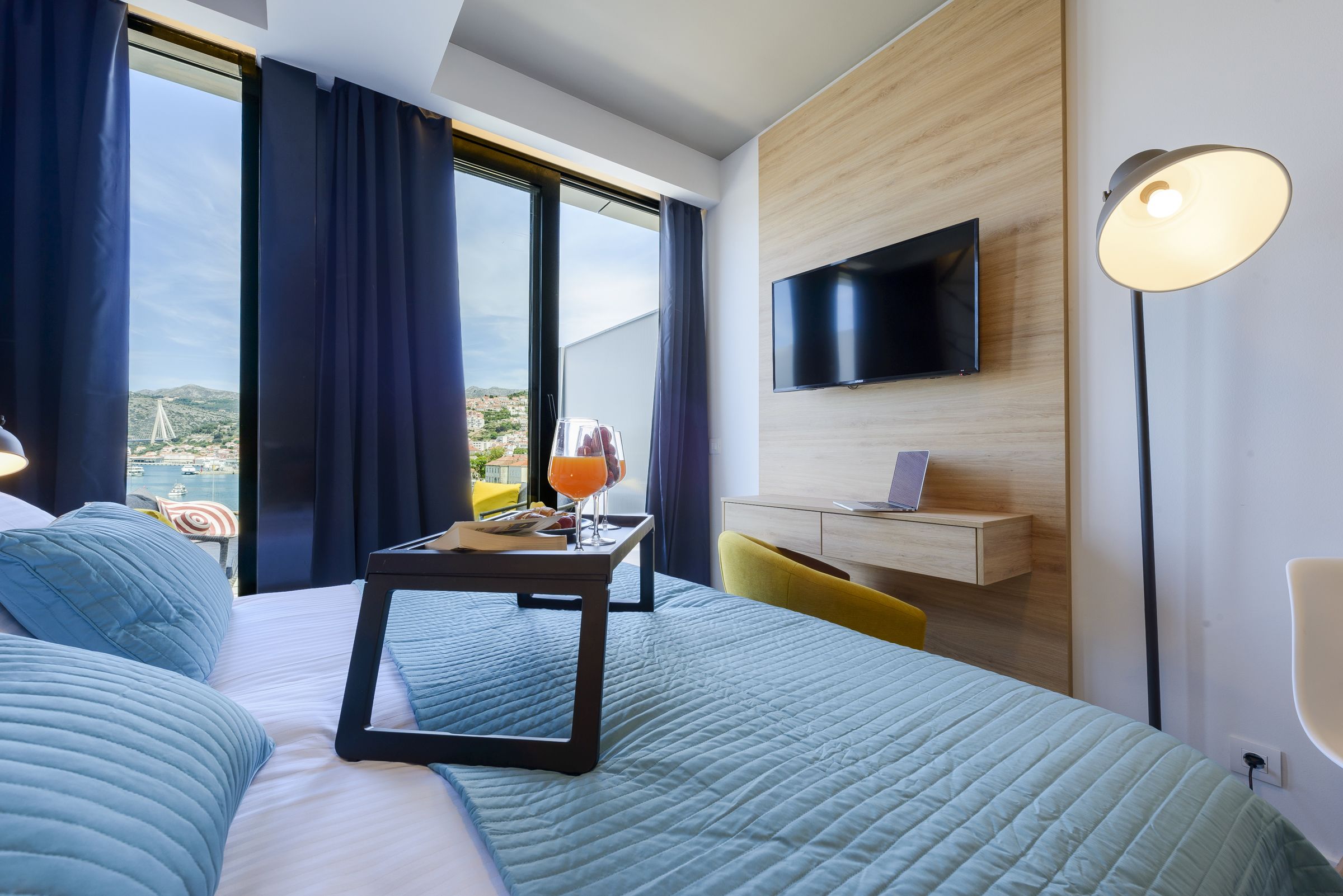 A hotel room with a bed, a desk with a laptop, a TV mounted on the wall, and a view of a bridge and landscape.