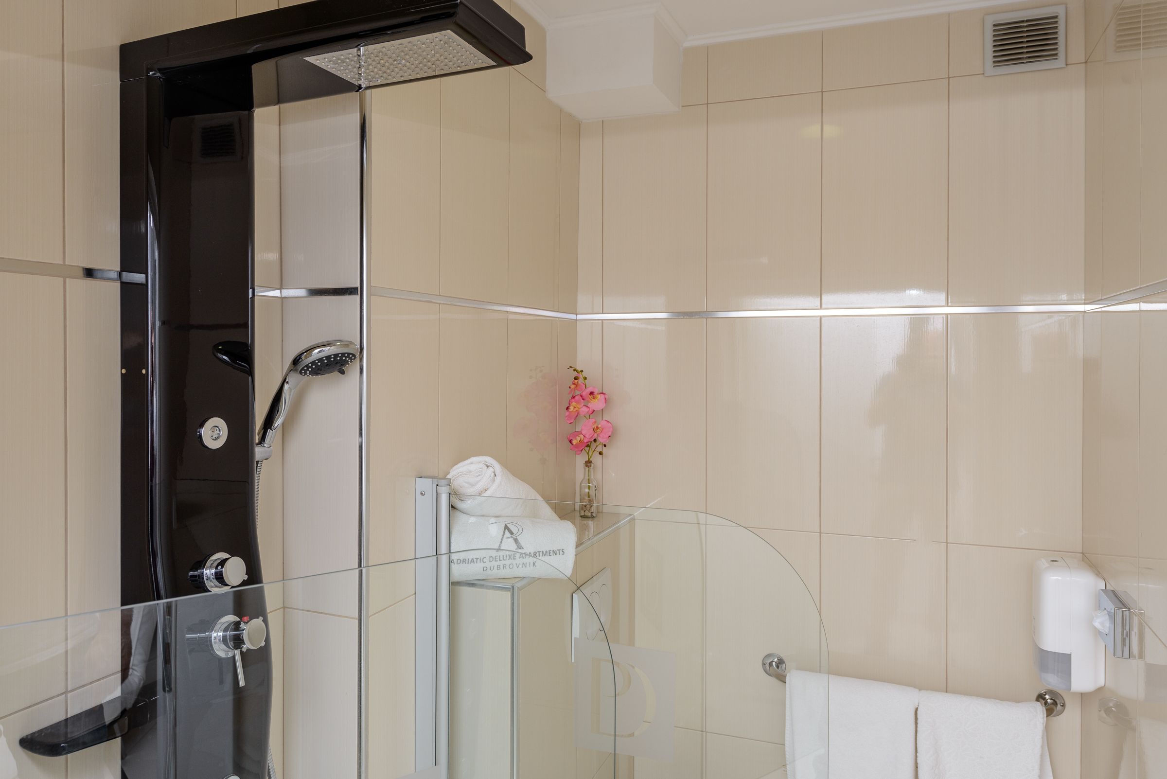 A modern bathroom with a glass-enclosed shower, a towel rack with a white towel, and a small vase with pink flowers on the wall.