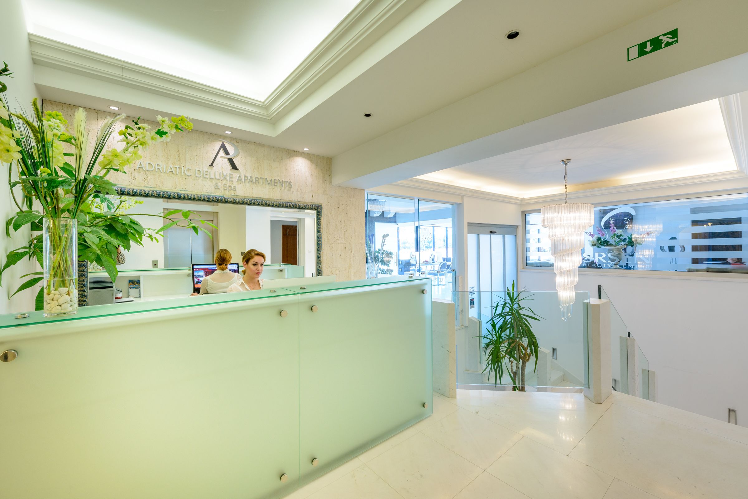 Un espace d’accueil d’hôtel avec un comptoir en verre, un bureau d’accueil et une enseigne indiquant « Adriatic Deluxe Apartments ».
