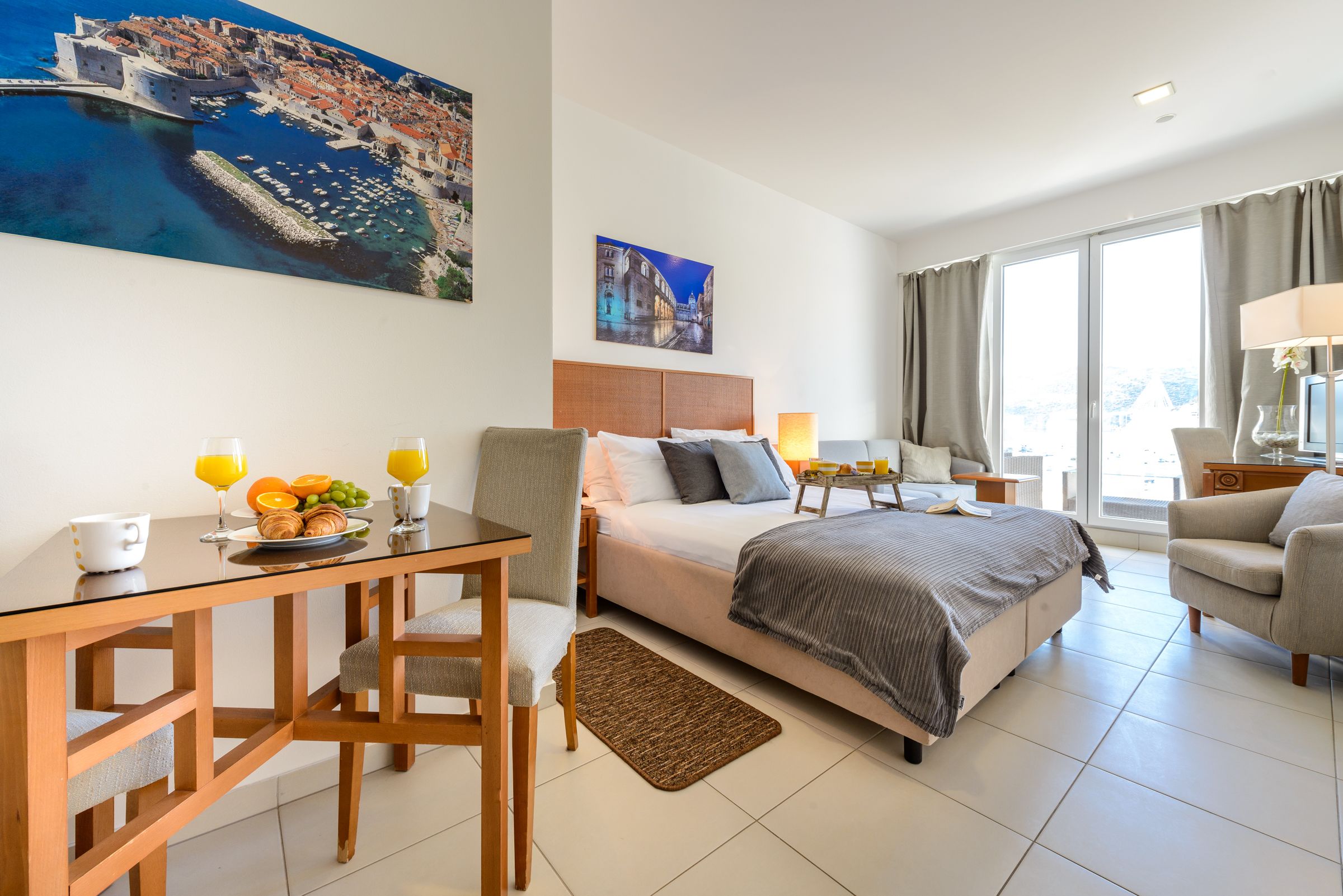 A hotel room with a bed, a desk with breakfast, a chair, a lamp, and a painting of a coastal town.