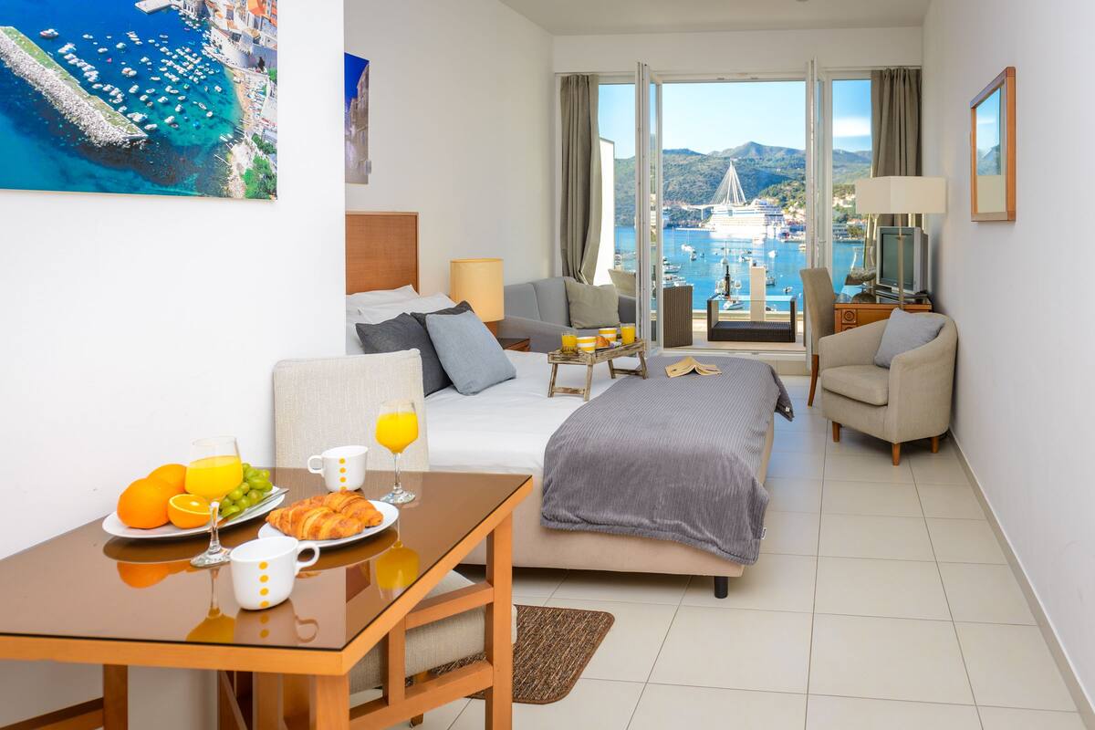 Une chambre d’hôtel avec un lit, un canapé, une table à manger où le petit-déjeuner est servi, et une vue sur la mer et les bateaux.