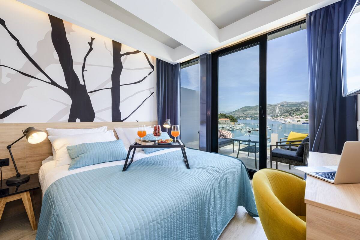 Une chambre d’hôtel moderne dotée d’un grand lit, d’un bureau avec un ordinateur portable, d’une table à manger avec un plateau de jus d’orange, et offrant une vue panoramique sur la ville et les montagnes.