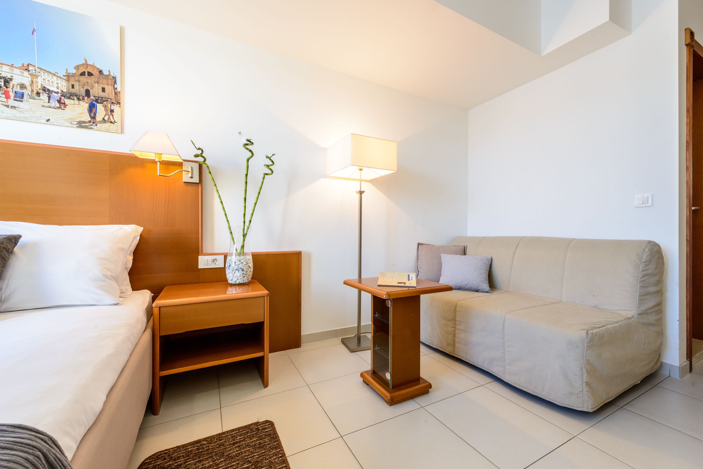 A hotel room with a bed, a sofa, a small wooden table, a floor lamp, and a wall-mounted picture.