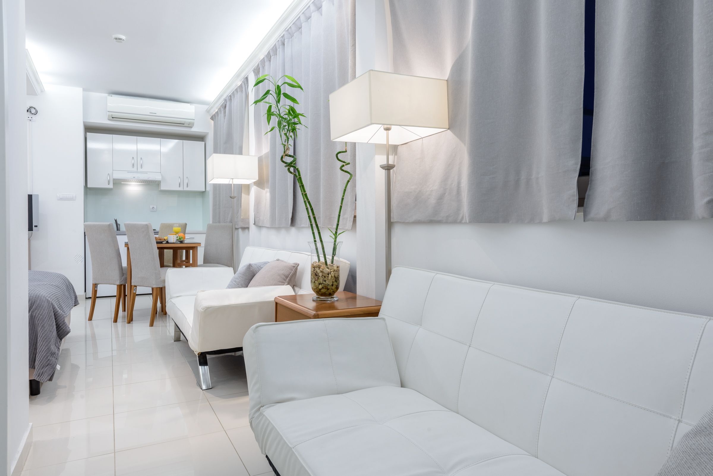 A modern, compact living space with a white sofa, a dining table with chairs, a small kitchen area, and a bedside table with a lamp and a plant.