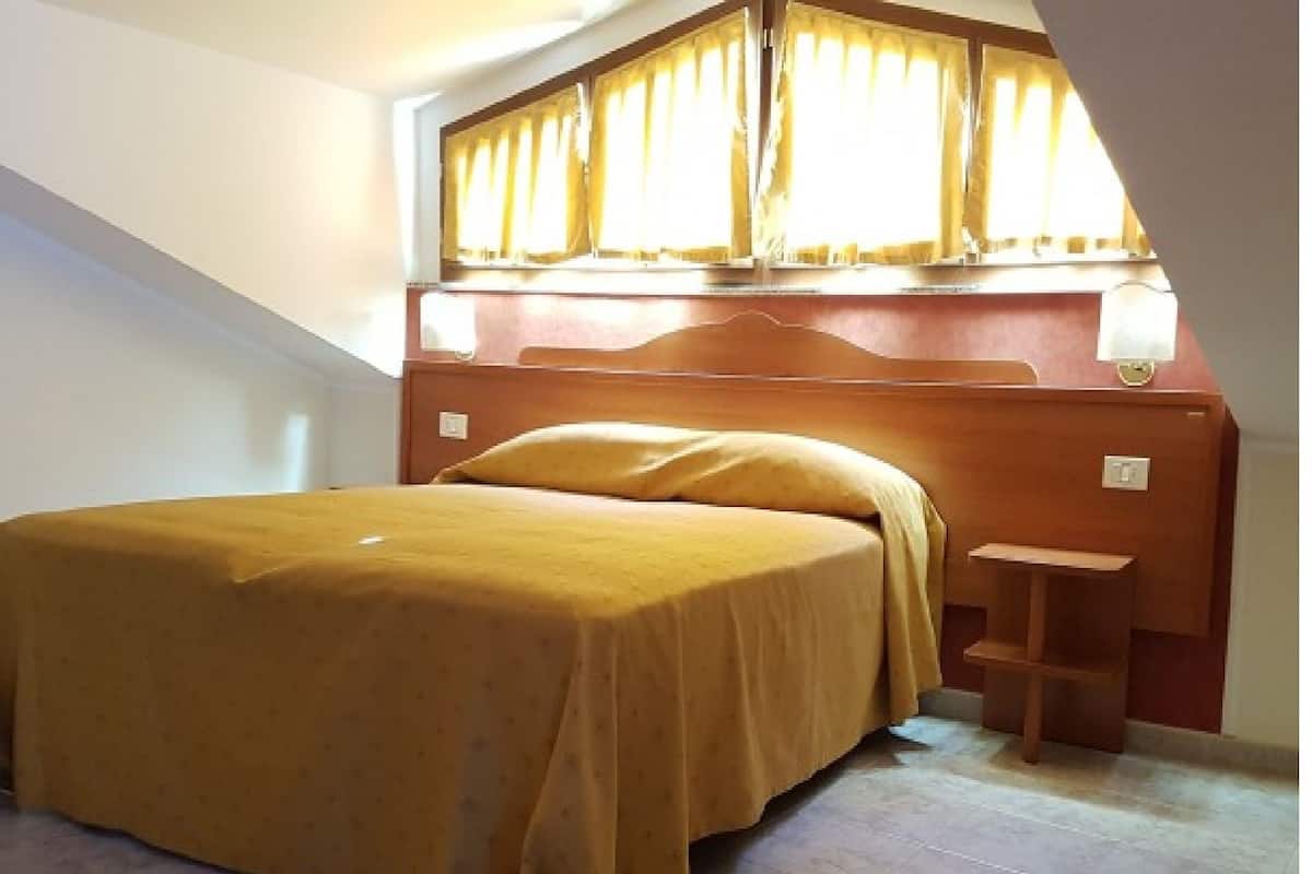 Een slaapkamer met een bed, een nachtkastje, een raam met gordijnen en een schuine plafond.