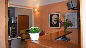 A reception area with a wooden reception desk, a small white table, a yellow sofa, a framed portrait, and a wall-mounted television. - Hotel dei Cappuccini (San Giovanni Rotondo)