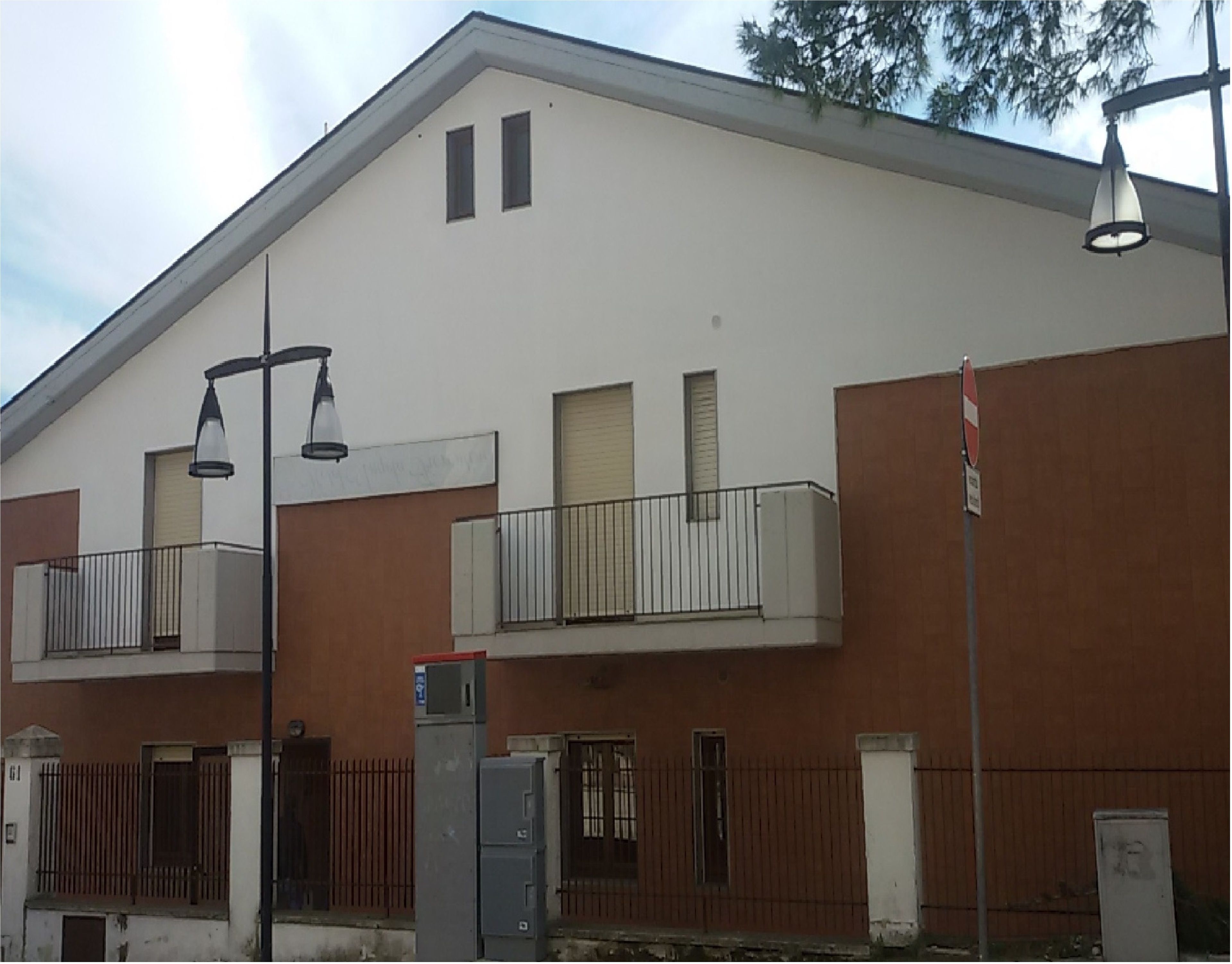 Un edificio de dos pisos con techo plano, un balcón en el segundo nivel y un portón de seguridad en la entrada.