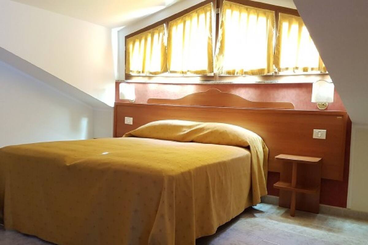Een slaapkamer met een bed, nachtkastje, raam met gordijnen en een schuin dak.