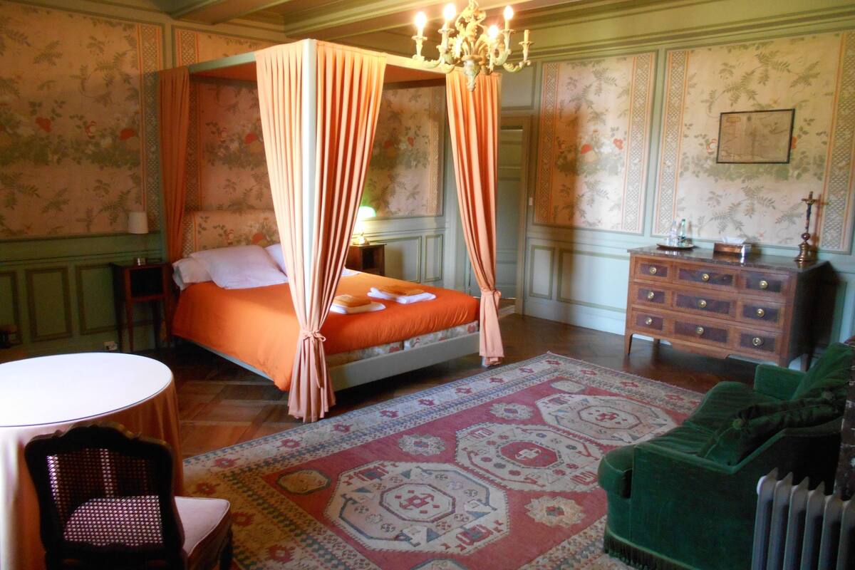 A four-poster bed with a canopy, a green sofa, a wooden dresser, and a patterned rug in a room with floral wallpaper.