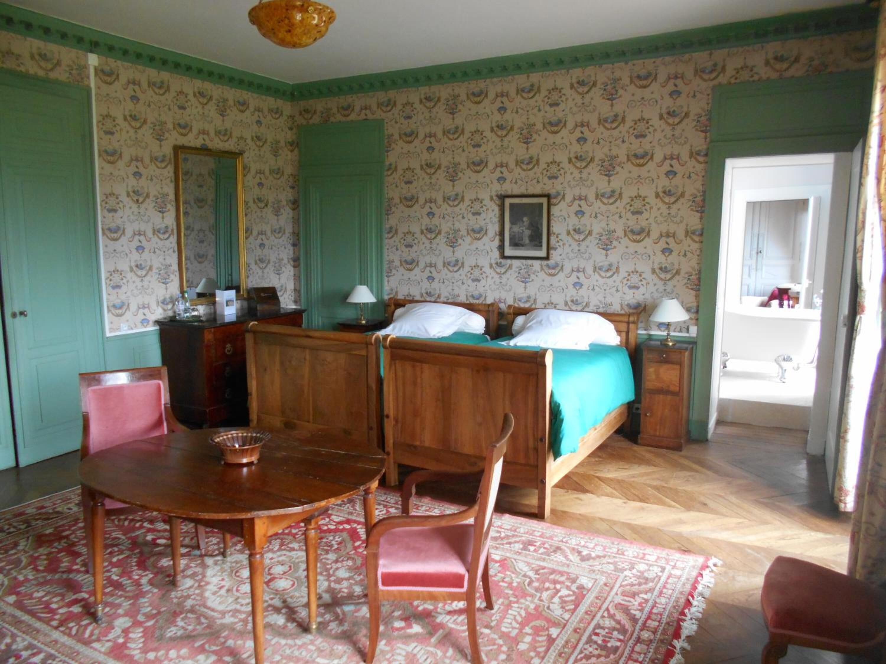 A bedroom with a wooden bed, a bedside table, a chair, a table with a bowl, patterned wallpaper, and a door leading to a bathroom.