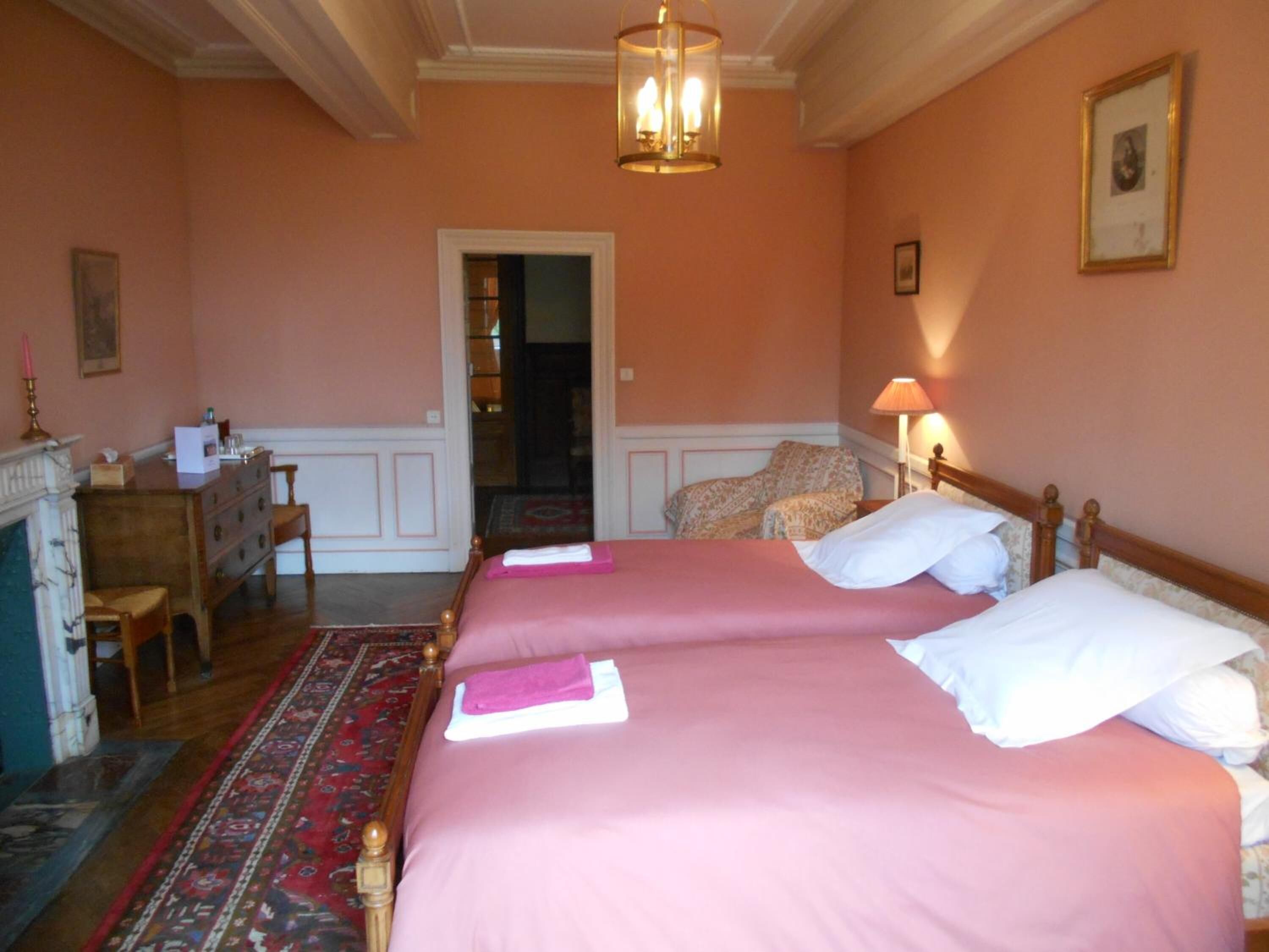 A hotel room with two beds, a desk, a chair, a fireplace, and a chandelier.