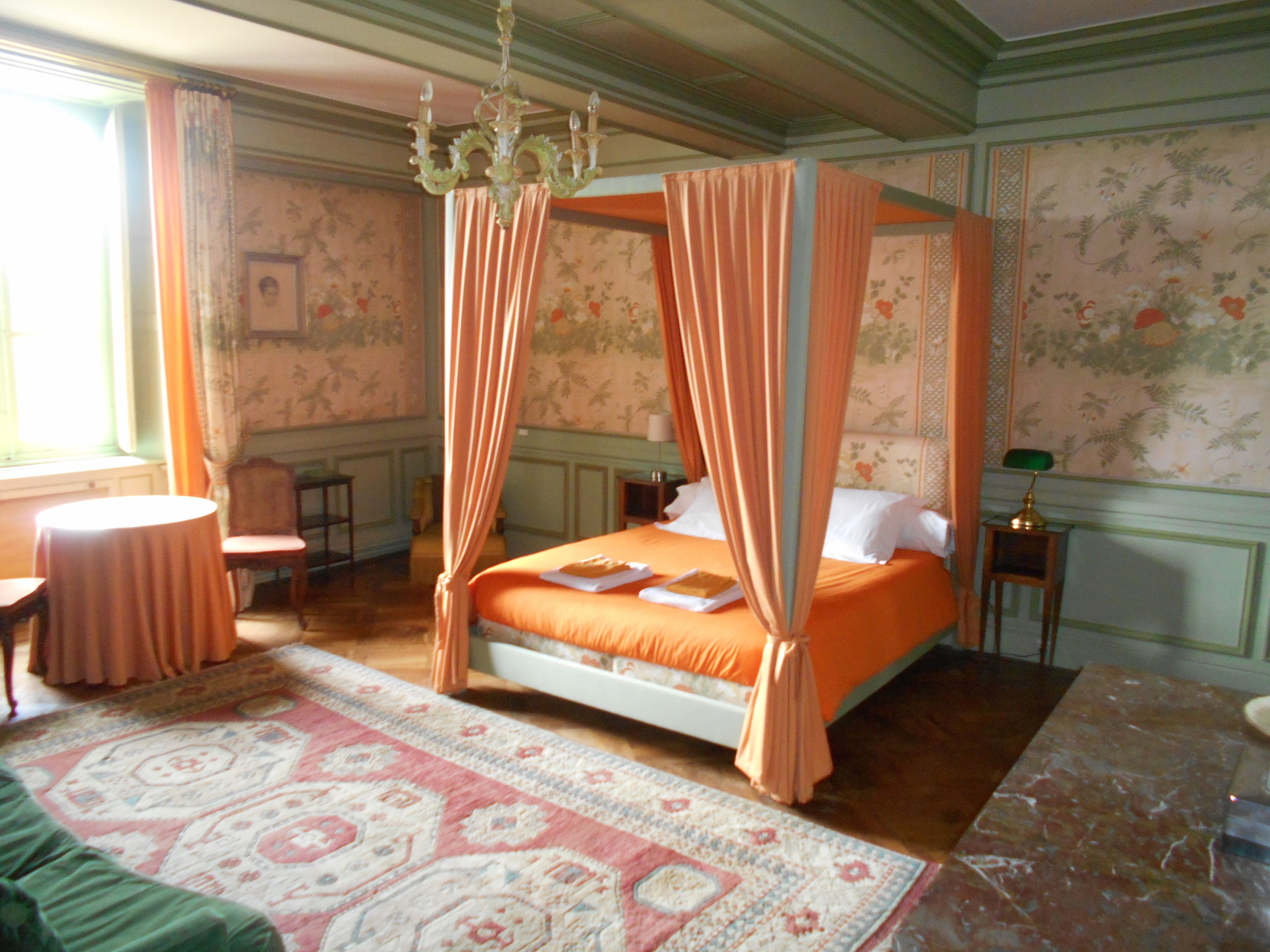 Una cama con dosel y cuatro columnas, papel tapiz con motivos florales, una mesa redonda y un sofá verde.