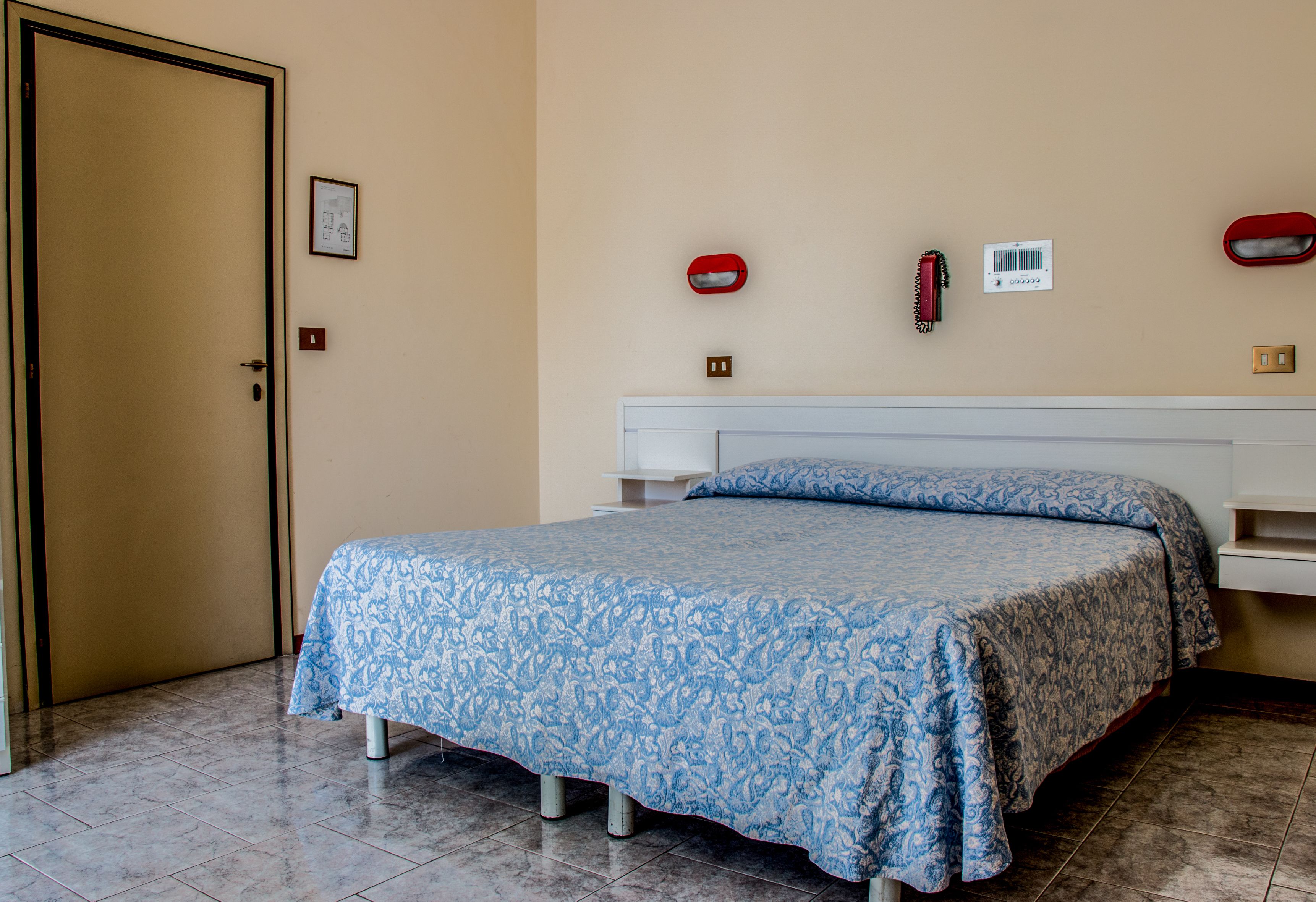 Een slaapkamer met een bed, een deur, een aan de muur bevestigd telefoon en een thermostaat.