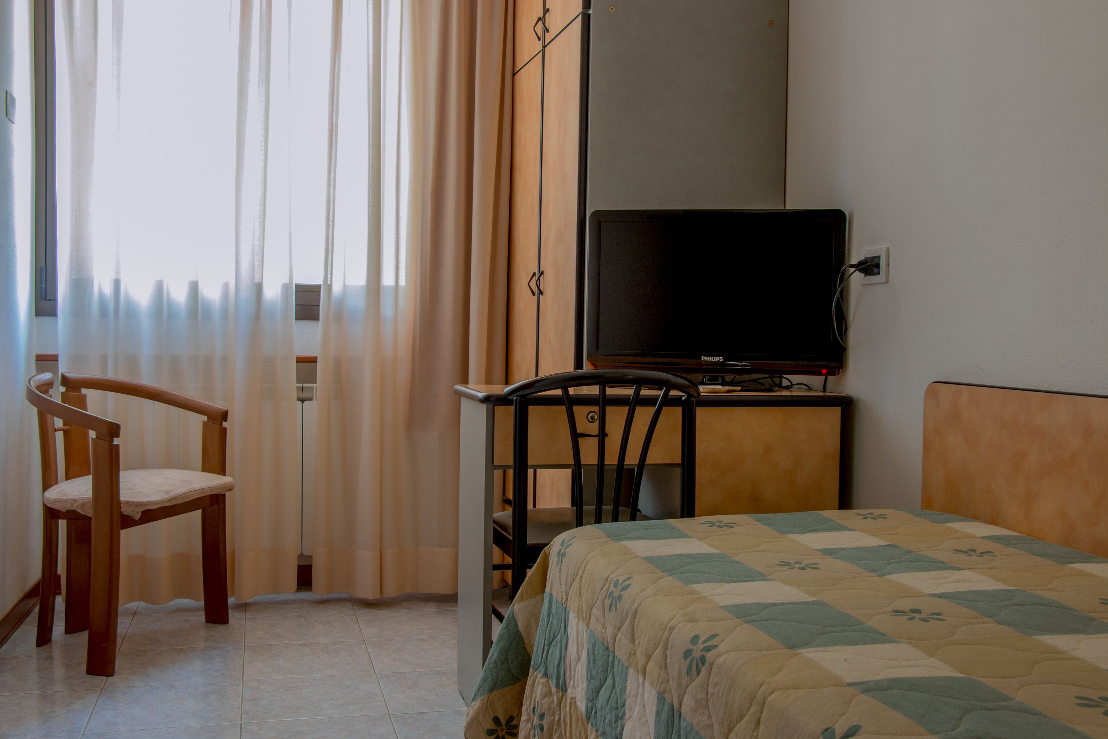 A room with a bed, a television on a stand, a wooden chair, and a window with curtains.