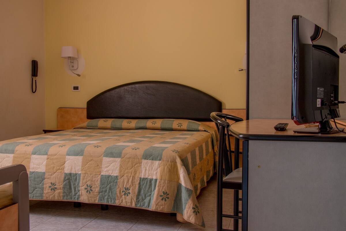 A hotel room with a bed, a desk with a computer, a chair, a lamp, and a phone on the wall.