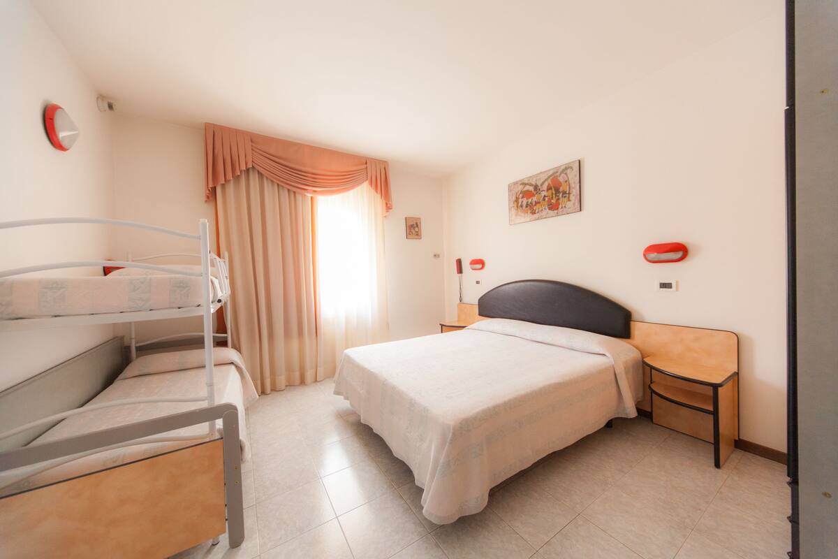 A hotel room with a bunk bed, a single bed, a nightstand, a wall clock, a painting, and a window with curtains.
