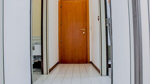Un pasillo estrecho con una puerta de madera al final, que da a una habitación con cama y mesita de noche.