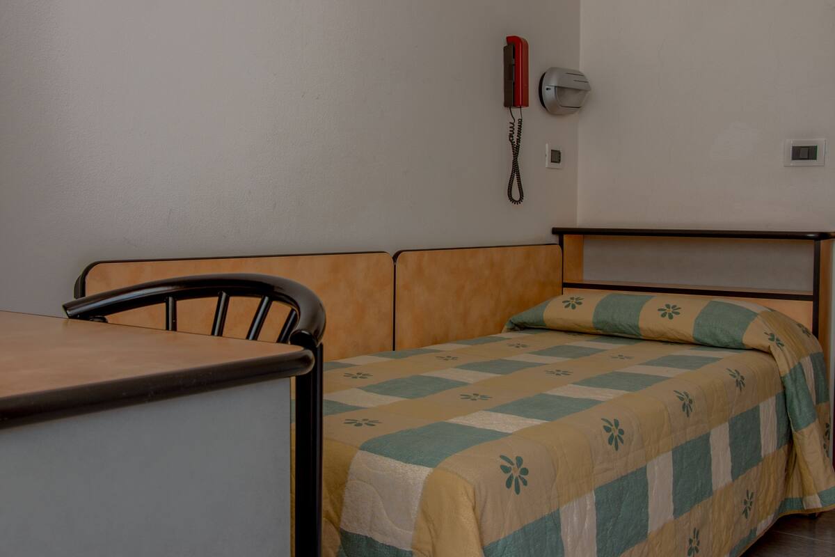 A single bed with a quilt, a chair, a bedside table, a telephone, and a wall-mounted light fixture.