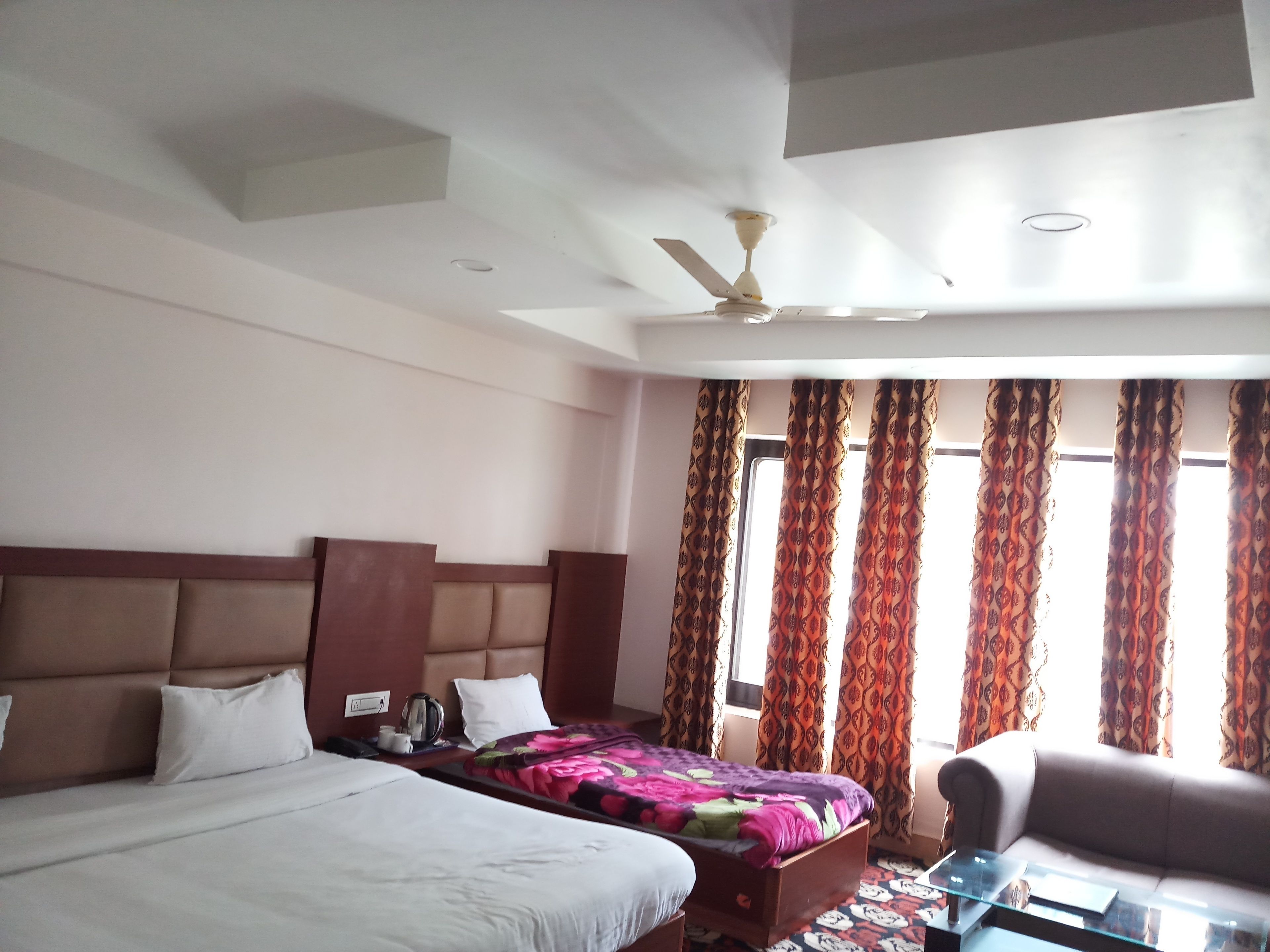 A hotel room with two beds, a sofa, a ceiling fan, and patterned curtains.