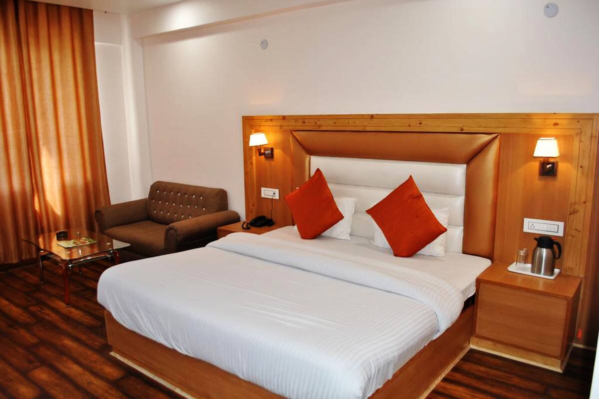 Quarto de hotel com uma cama grande, duas mesas de cabeceira, um painel de cama de madeira, um sofá marrom e uma mesa de centro de vidro.