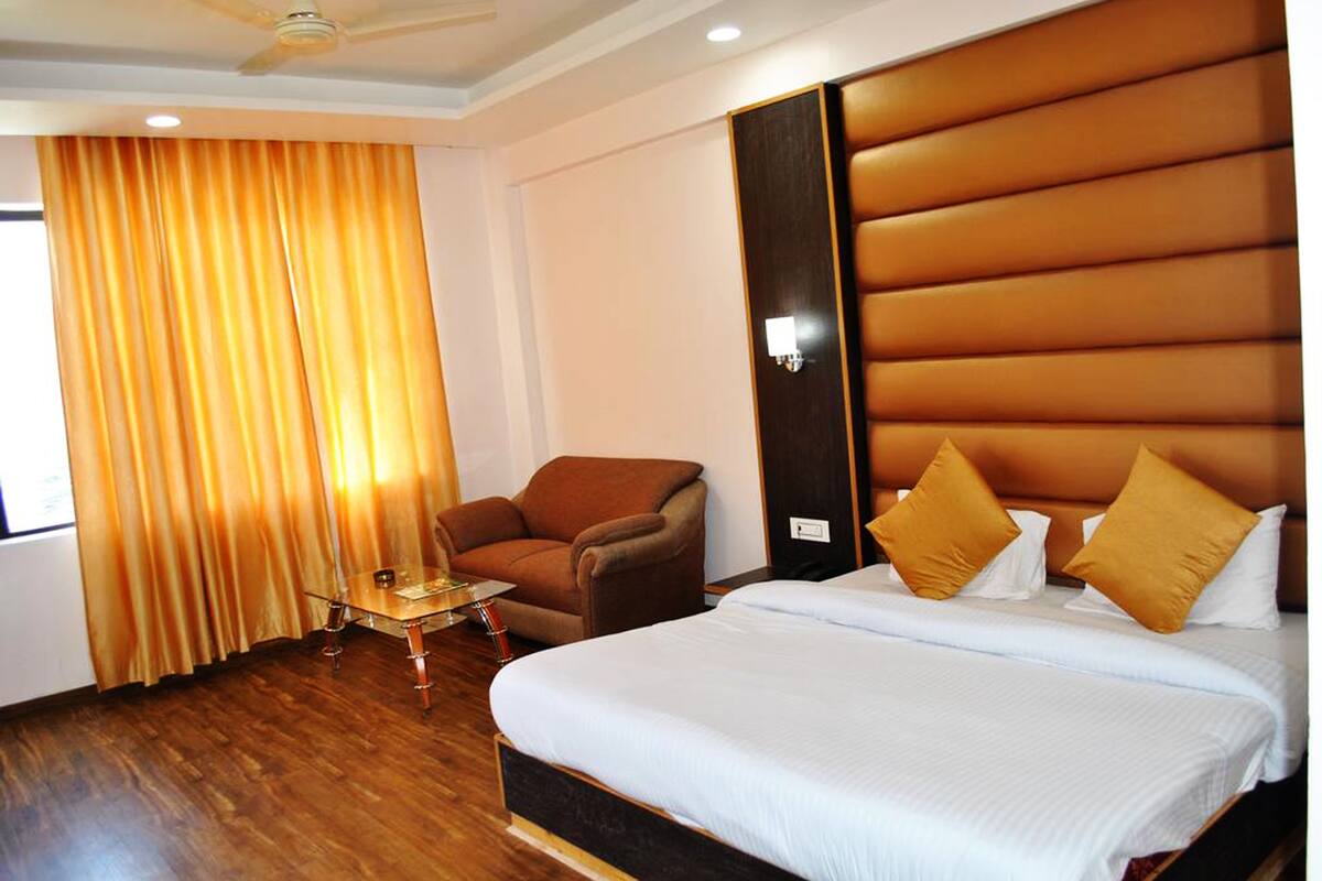 Quarto de hotel com uma cama grande, uma poltrona marrom, uma mesinha e uma cortina.