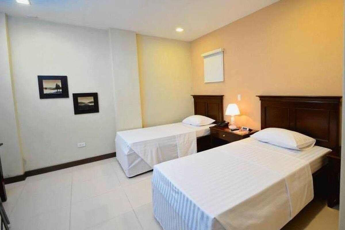 Una habitación de hotel con dos camas individuales, una mesita de noche con lámpara y dos cuadros en la pared.