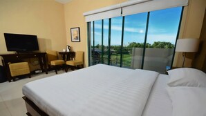 A hotel room with a large bed, a desk with a television, a chair, and a view of the outdoors through large windows.