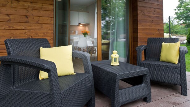 Un patio con dos sillas de mimbre y una pequeña mesa, una puerta de cristal que da a un espacio interior y una pared exterior de madera.