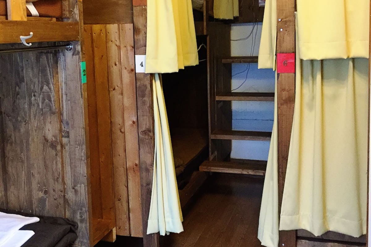 Ein schmaler Korridor mit Holzwänden und Holzboden. Gelbe Vorhänge hängen von der Decke, und ein Bett mit weißer Bettdecke ist auf der linken Seite zu sehen.