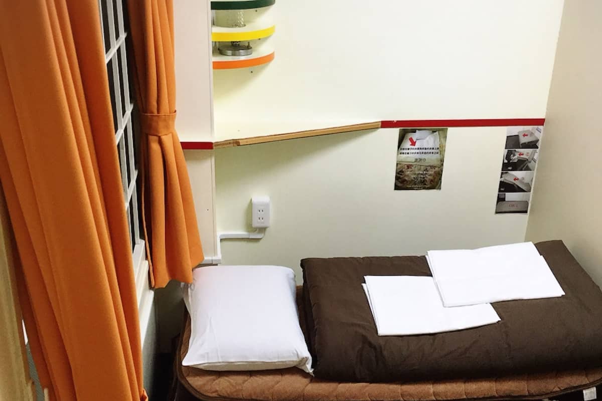 Ein kleines Zimmer mit einem Einzelbett, einer Matratze, Kissen und einem Regal mit diversen Gegenständen.