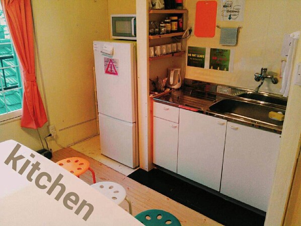 Cocina compacta con gabinetes blancos, microondas, fregadero y refrigerador. Hay varios utensilios y electrodomésticos en las encimeras.