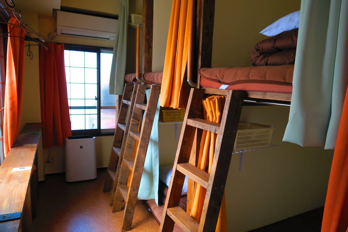 Ein Schlafraum mit Etagenbetten, einer Leiter, einem Fenster mit Vorhängen und einer kleinen Klimaanlage.
