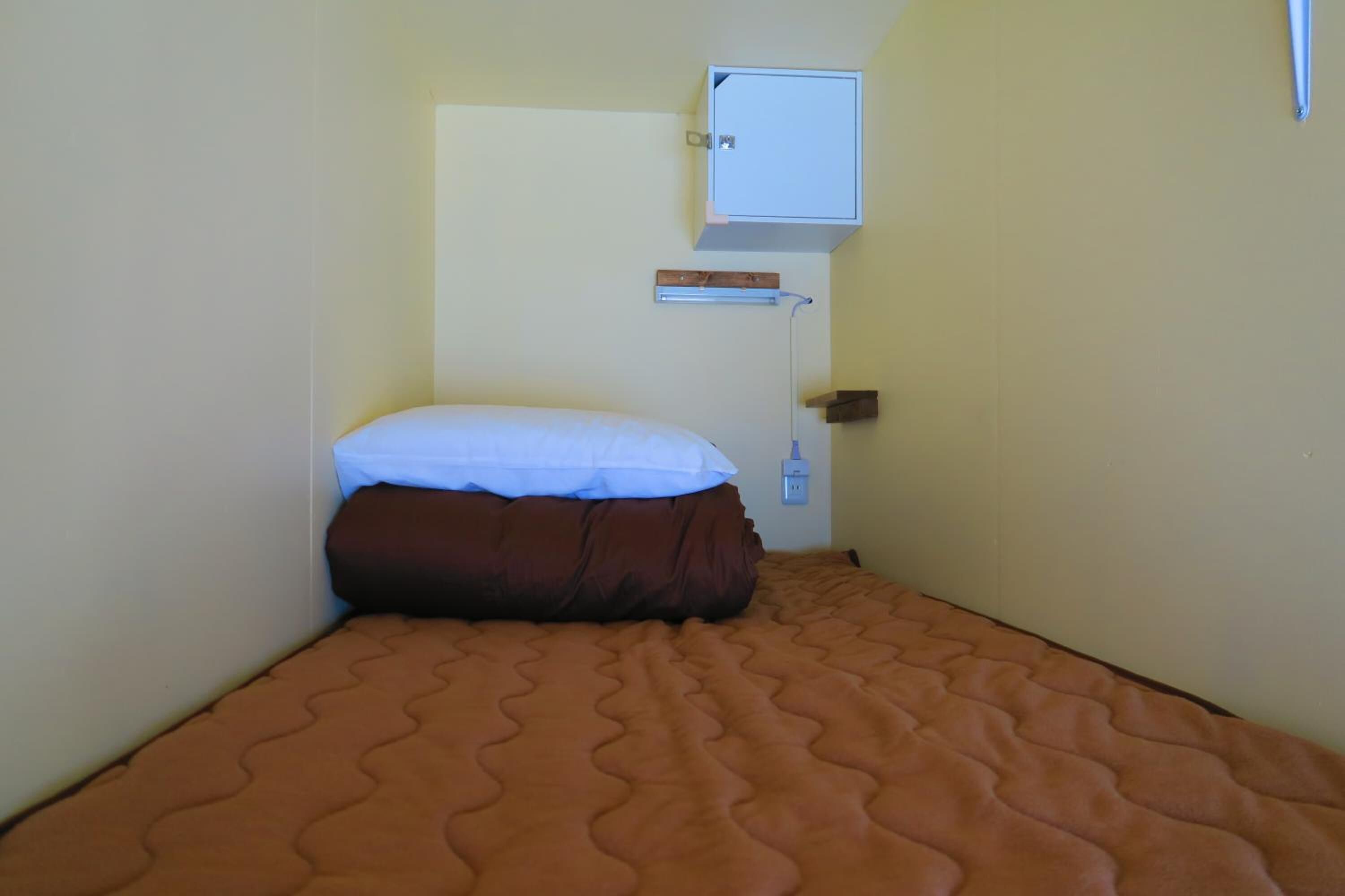 Una habitación pequeña con cama individual, colchón marrón, almohada blanca y un mueble alto de color blanco sobre la cama.